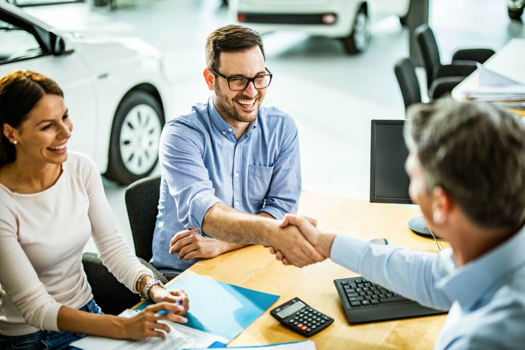 Happy couple buying a car in a showroom while men are shaking hands after reaching a successful deal. Focus is on man with glasses.